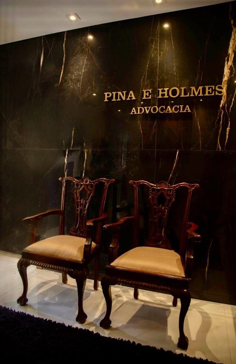 Recepção do escritório Pina e Holmes Advocacia, com o nome do escritório em letras douradas sobre parede de mármore escuro e duas cadeiras de madeira entalhada